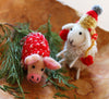 Two felted animals, a sheep and a pig, wearing a festive hat and blanket on a wooden surface with greenery.