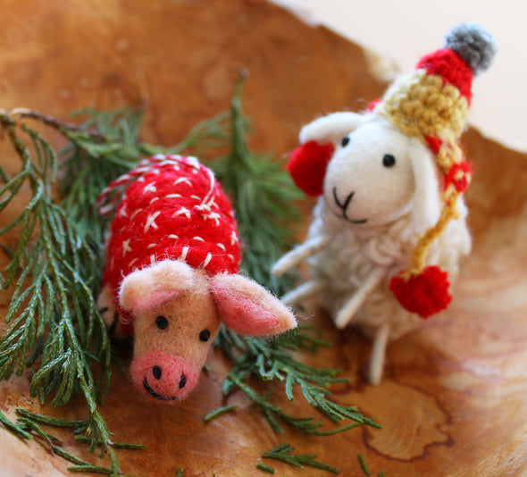 Two felted animals, a sheep and a pig, wearing a festive hat and blanket on a wooden surface with greenery.