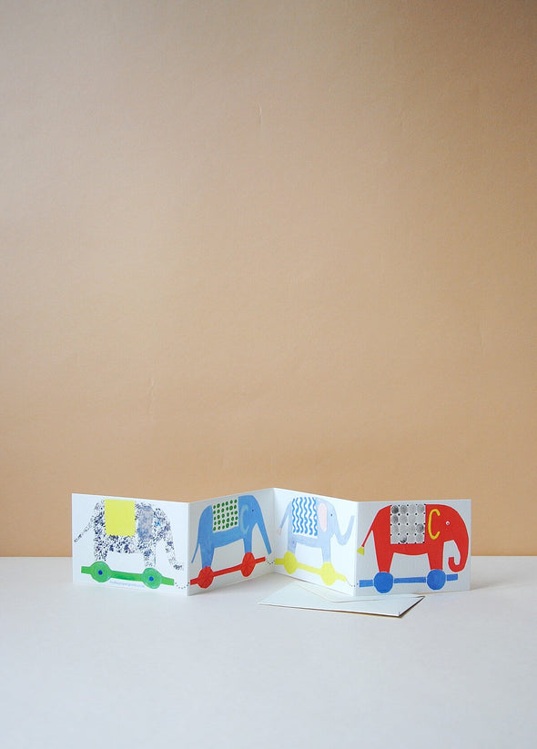 Greetings card depicting four toy elephants in a row. Each elephant is a different colour and is on contrasting coloured wheels.