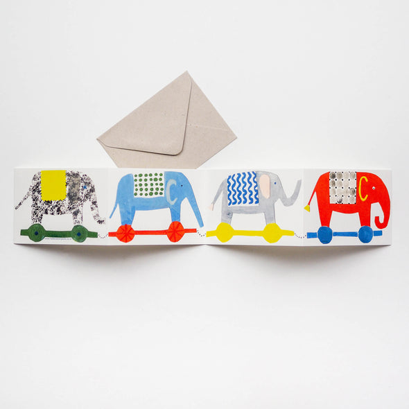 Greetings card depicting four toy elephants in a row. Each elephant is a different colour and is on contrasting coloured wheels.