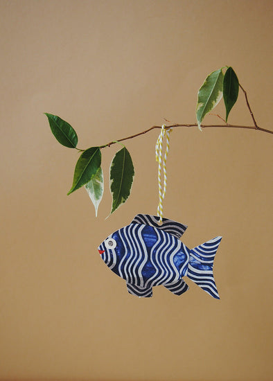 A gift tag in the shape of a fish with a loop of string attached. The fish is blue with white wavy stripes and a red smiling mouth. The gift tag is hanging from a thin branch which has a few green leaves sprouting off it.