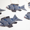 Close up of gift tags in the shape of fish with a loop of string attached to each. The fish are blue with white wavy stripes and a red smiling mouth.