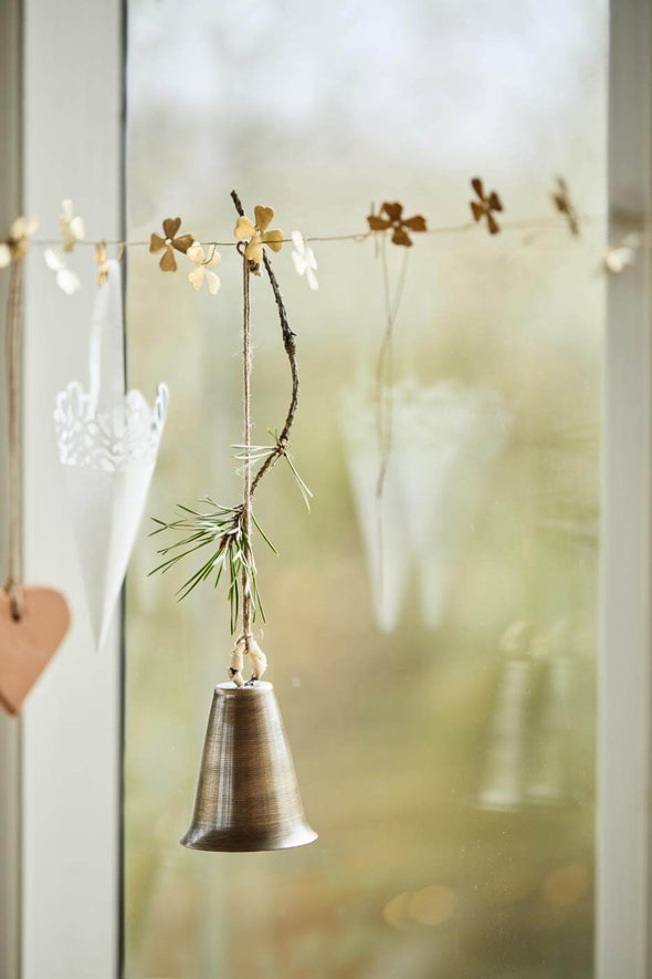 A garland of small brass flowers strung across a window with a brass bell hanging from them.