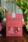 A pink paper packet containing a paper garland. The packet is resting upright against a tray of cupcakes, with flowers in the background.