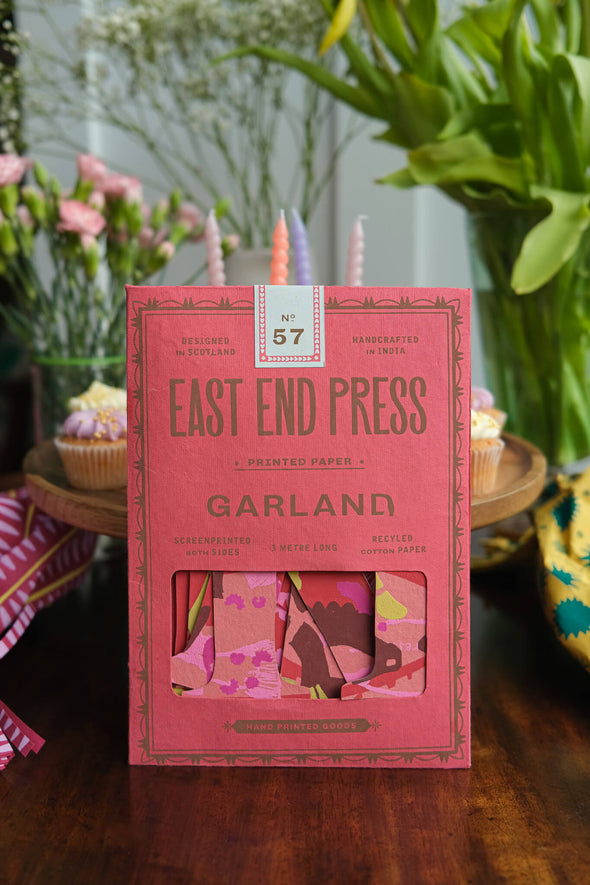 A pink paper packet containing a paper garland. The packet is resting upright against a tray of cupcakes, with flowers in the background.