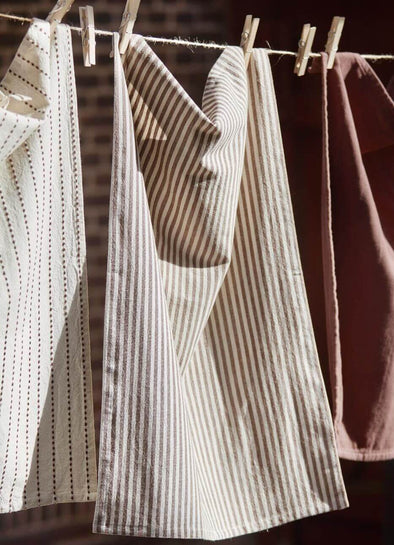 Three tea towels hanging from a washing line in the sun. The towel on the left is cream with a brown interwoven stripe, the middle has cream and brown stripes and the towel on the right is brown coloured.