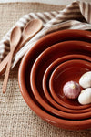 A nest of four terracotta coloured ceramic bowls containing a few garlic bulbs. Next to the bowl lies a cream and brown striped tea towel on which lies a pair of wooden salad servers.
