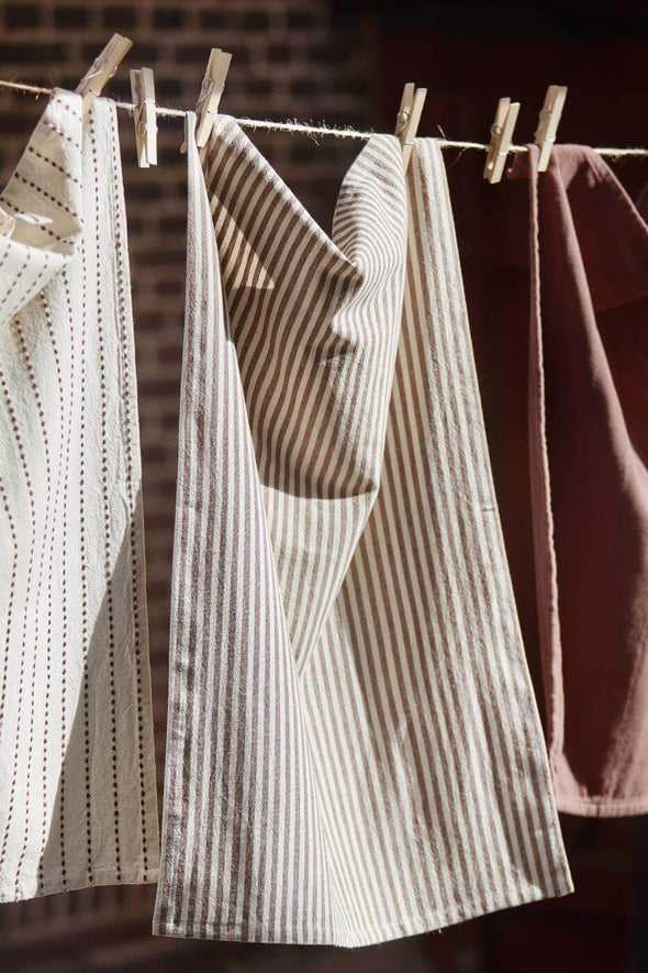 Three tea towels hanging from a washing line in the sun. The towel on the left is cream with a brown interwoven stripe, the middle has cream and brown stripes and the towel on the right is brown coloured.