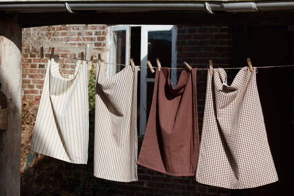 Four tea towels hanging from a washing line in the sun. The towel on the far left is cream with a brown interwoven stripe, the next has cream and brown stripes, the third is solid brown and the last is a cream and brown checked pattern.