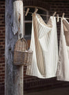 Two tea towels hanging from a washing line in the sun. The towel on the left is cream with a brown interwoven stripe, the other has cream and brown stripes. A basket full of pegs hangs from a wooden post next to the washing line.