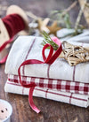 A pile of three kitchen towels, the bottom is cream with a big red checked pattern, the middle is cream and red gingham and the top is cream with a thin red stripe. Some Christmas decorations and red ribbon are strewn around.