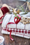 A pile of three kitchen towels, the bottom is cream with a big red checked pattern, the middle is cream and red gingham and the top is cream with a thin red stripe. Some Christmas decorations and red ribbon are strewn around.