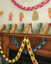 Yellow and blue paper chains looped across a wooden mantelpiece. A paper garland of stockings rests along the top of the mantelpiece and a red paper chain hangs on the wall above.