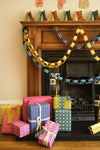 Black fireplace and wooden mantelpiece surrounded by Christmas decorations. Yellow and blue paper chains hang from the mantelpiece. Colourful presents are piled on the ground in front of the fireplace.