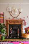 Black fireplace and wooden mantelpiece surrounded by Christmas decorations. A red and pink paper chain trails across the mantelpiece and colourful paper garlands of baubles hang above and below. On the white wall above the fireplace hang three large paper snowflakes with a multi coloured paper chain draped between them.