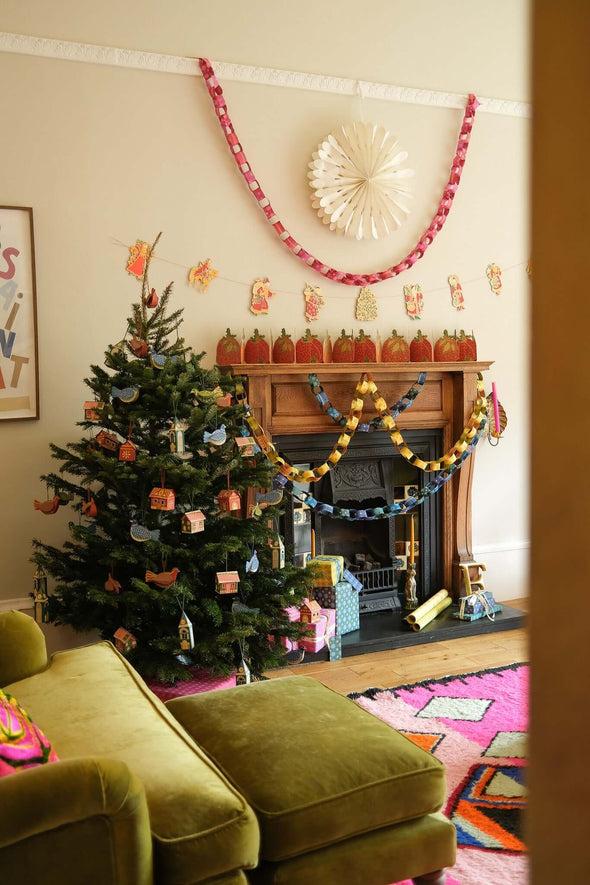 Black fireplace and wooden mantelpiece surrounded by Christmas decorations. Yellow and blue paper chains hang from the mantelpiece. On the white wall above the fireplace hangs a large paper snowflake with a red and pink paper chain draped around it. A decorated Christmas tree stands next to the fireplace.