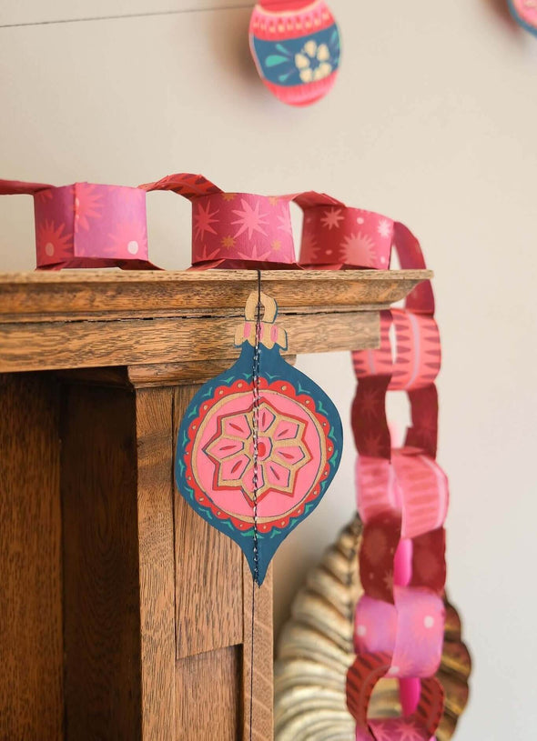 Red and pink paper chain trailing over the corner of a wooden mantelpiece. A colourful, paper bauble hangs down from the mantelpiece just in front.