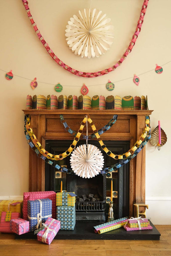 Black fireplace and wooden mantelpiece surrounded by Christmas decorations. Yellow and blue paper chains hang from the mantelpiece. On the white wall above the fireplace hangs a large paper snowflake with a red and pink paper chain draped around it. Colourful presents are piled on the ground in front of the fireplace.