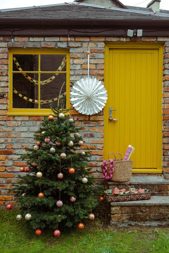 Brick cottage with a yellow door and a square window with a yellow painted frame. A Christmas tree decorated with baubles stands by the door. A yellow paper chain can be seen strung across the inside of the window.