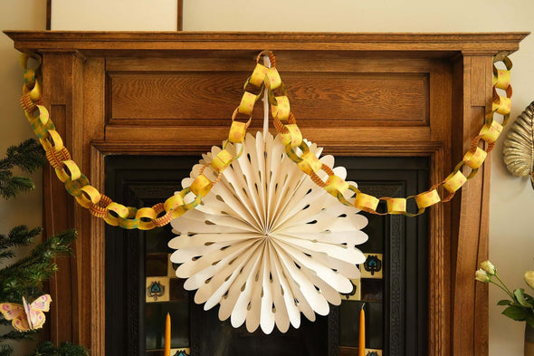 A giant paper snowflake hanging in front of a black fireplace and wooden mantelpiece. A yellow and green paper chain is strung across the mantelpiece hanging down in two loops.