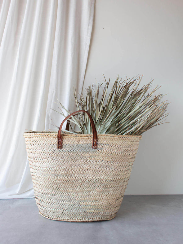Basket bag with short tan leather handles on a grey floor and white fabric background. Palm leaves rest in the bag.