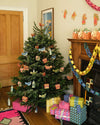 A decorated Christmas tree standing next to a fireplace. The tree is covered in hanging decorations made from paper board. They are a mix of red gingerbread houses, green steeples, red robins, blue doves and green partridges. Lots of colourfully wrapped gifts are piled under the tree.