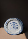 Ceramic plate with a white glaze and cobalt blue painted illustration of three sardines. The plate is sitting in a pool of light.