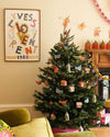 A decorated Christmas tree standing next to a fireplace. The tree is covered in hanging decorations made from paper board. They are a mix of red gingerbread houses, green steeples, red robins, blue doves and green partridges.