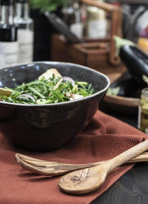 Pair of wooden salad servers lying in front of a bowl of salad on a rust coloured tea towel.