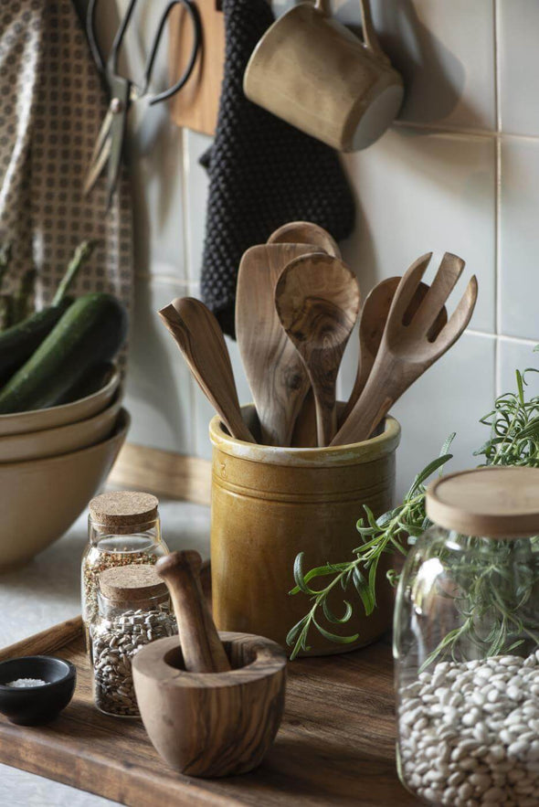 A mustard coloured ceramic utensil pot filled with wooden utensils. Some glass jars filled with beans and a wooden pestle and mortar sit to the front.
