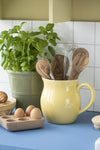 A yellow ceramic jug filled with wooden utensils and a metal whisk. A wooden egg holder filled with three eggs sits alongside and a green ceramic pot filled with a basil plant is just behind.