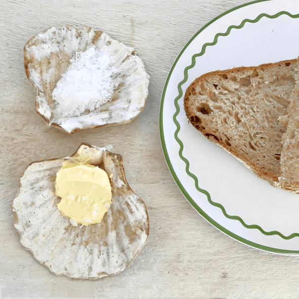 Two ceramic scallop shells glazed in cream and light brown. One is holding some salt, the other a knob of butter. A white and green plate with some toast resting on it, is next to the shells.