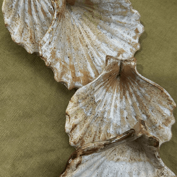 Close up of four ceramic scallop shells glazed in cream and light brown. They are resting on a green material.
