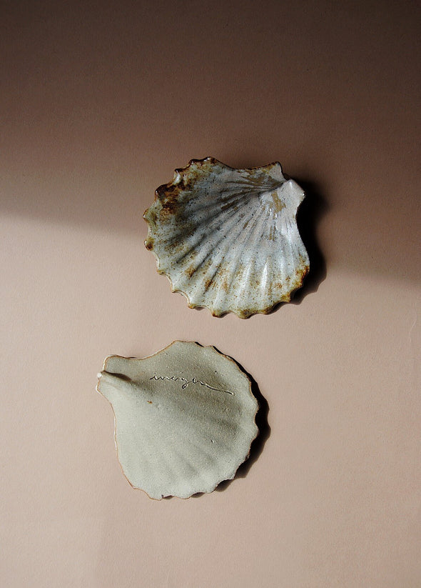 Two ceramic scallop shells glazed in cream and light brown. One shell is upside down showing its unglazed side with the artist's signature etched into the clay.