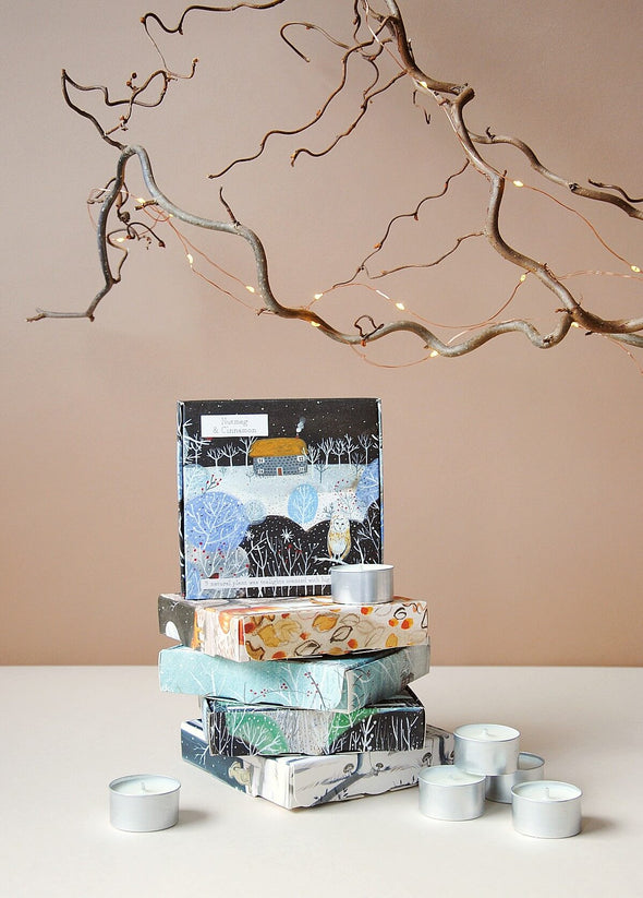 A pile of illustrated boxes. The top box sits upright and is illustrated with a winter scene showing snowy fields and trees with a stone cottage on the horizon and a white owl in the foreground. Tea lights  are scattered around the pile of boxes. Above the pile are some curly branches interwoven with tiny fairylights.