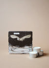 Square box illustrated with a starry sky and white snowy hills and trees. A large white owl swoops through the sky. Three tea lights sit next to the box.