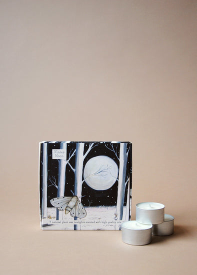 A square box illustrated with a large white moon in a starry sky and a pretty moth flying in front of some trees. Three tea lights sit next to the box.