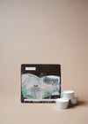 Square box illustrated with a winter scene depicting snowy hills and trees, two stone cottages and a red fox on the snowy hillside. Three tea lights sit next to the box.