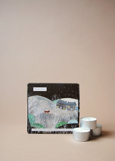 Square box illustrated with a winter scene depicting snowy hills and trees, two stone cottages and a red fox on the snowy hillside. Three tea lights sit next to the box.