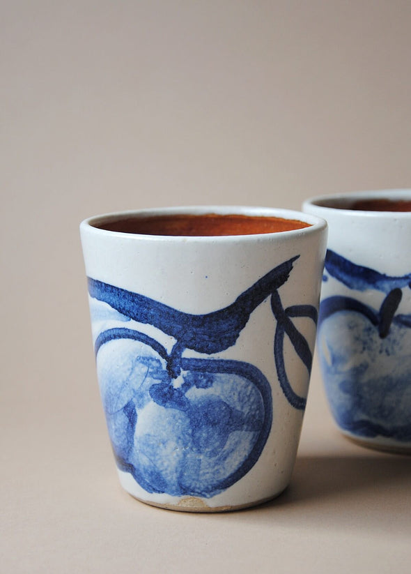 Ceramic tumbler in a white glaze with a blue hand painted design of apples.