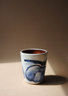 Ceramic tumbler in a white glaze with a blue hand painted design of apples. The tumbler is sitting in a pool of light.