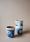 Three ceramic tumblers in a white glaze with a blue hand painted design of apples. Two tumblers are stacked, the other sits alongside.