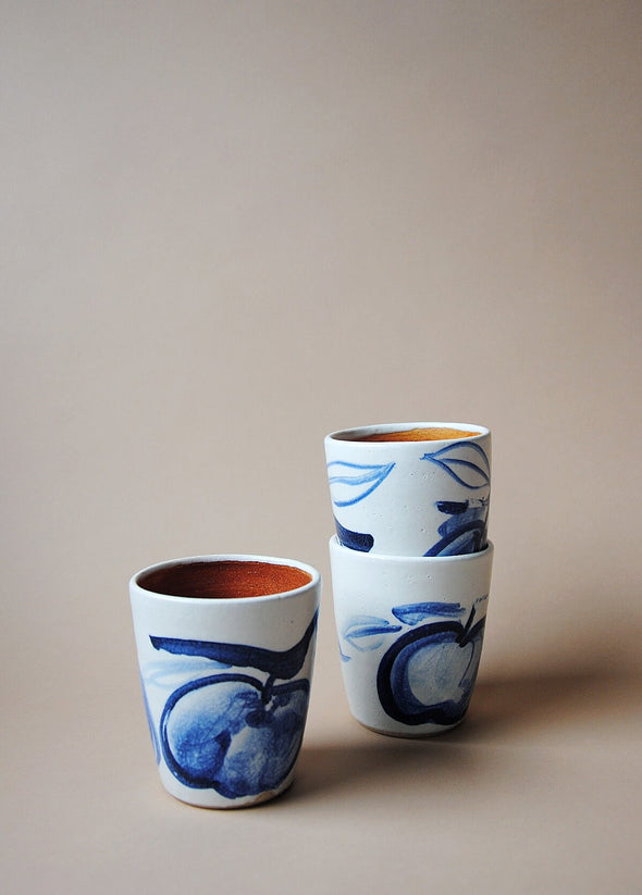 Three ceramic tumblers in a white glaze with a blue hand painted design of apples. Two tumblers are stacked, the other sits alongside.