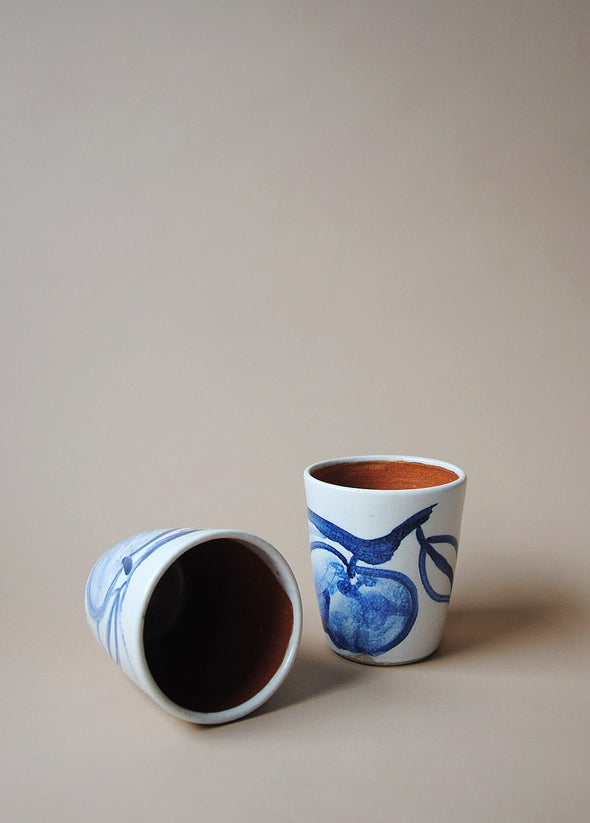 Two ceramic tumblers in a white glaze with a blue hand painted design of apples. One tumbler lies on its side.