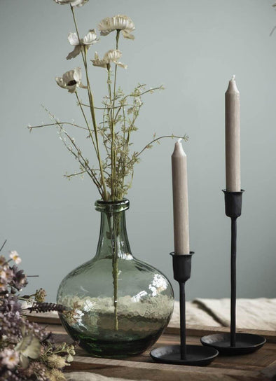 Green glass balloon shaped vase displaying a few dried stems. It sits on a wooden tray alongside two black candlesticks holding sand coloured tall dinner candles. 