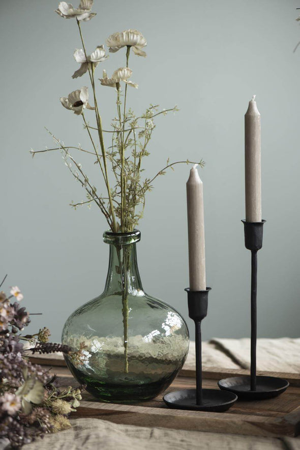 Green glass balloon shaped vase displaying a few dried stems. It sits on a wooden tray alongside two black candlesticks holding sand coloured tall dinner candles. 