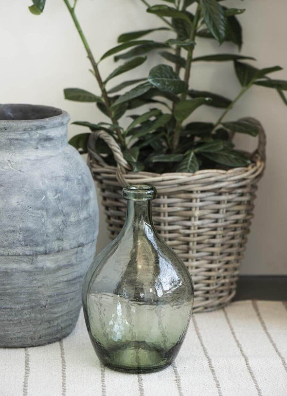 A green glass vase in a teardrop shape sitting in front of a basket containing a green plant and a grey ceramic large planter. 