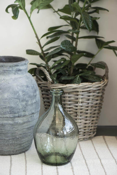 A green glass vase in a teardrop shape sitting in front of a basket containing a green plant and a grey ceramic large planter. 