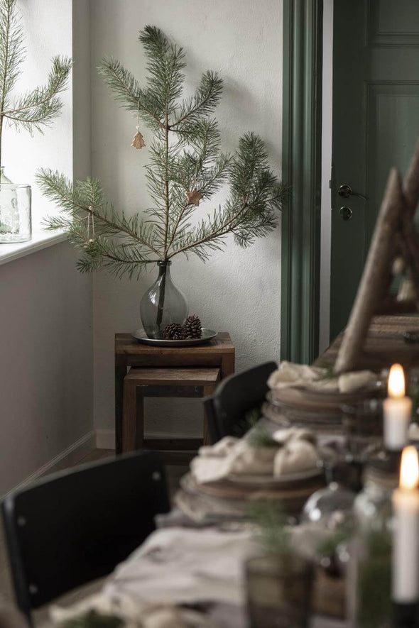 A grey glass teardrop shaped vase sitting on a small wooden table. The vase contains a single, large fir pine branch from which hangs a few Christmas decorations.