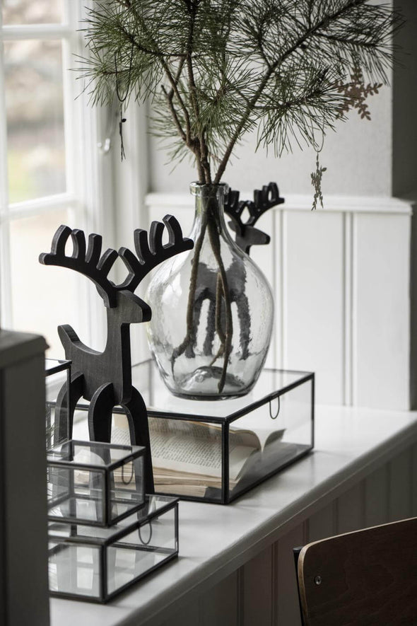 A grey glass teardrop shaped vase containing a few fir pine branches. Some Christmas decorations hang from the branches. Either side of the vase are two black wooden reindeer ornaments. 